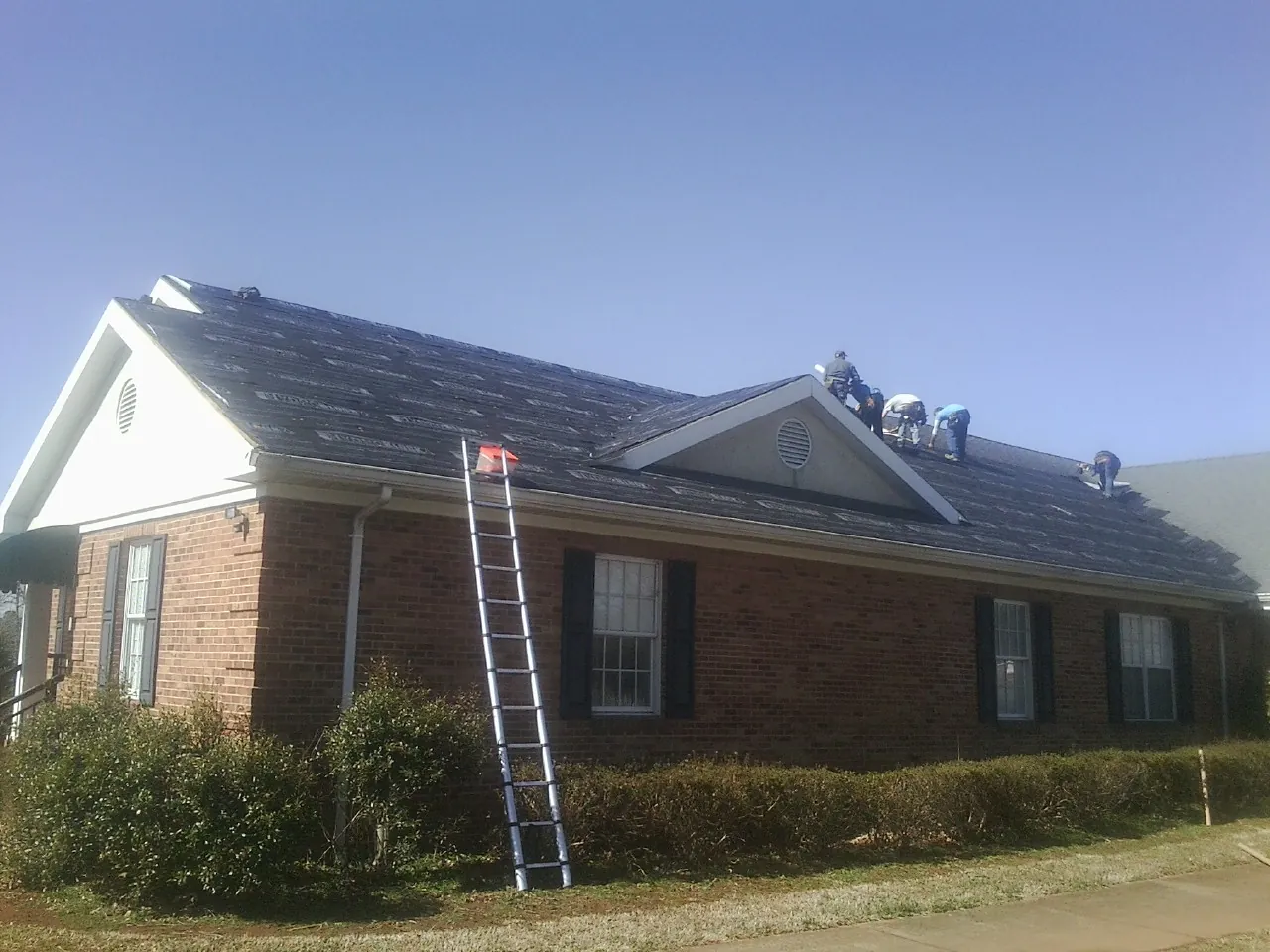 Crew tearing shingles off a brick ranch roof with a ladder set — Emergency Roof Repair work by our crew in Corapeake