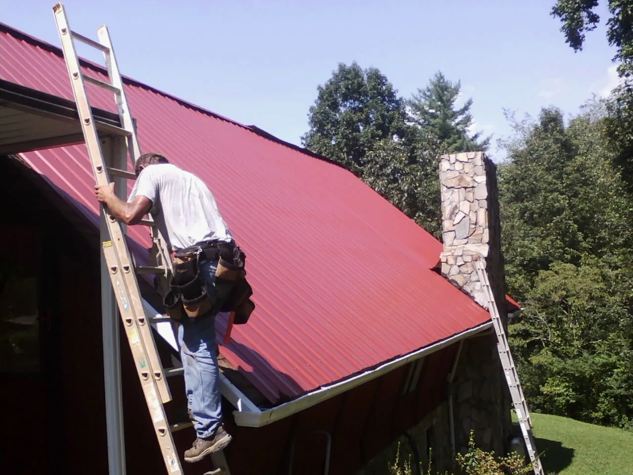 Roofer on a ladder beside a red metal roof and stone chimney — Soffit & Fascia Repair by Roofing Brothers in Corapeake
